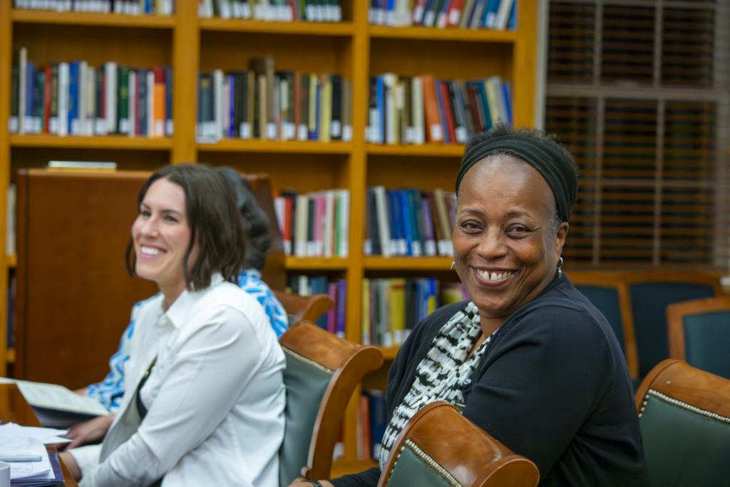 Black female teacher and white female teacher smiling and sitting at a table together. Window and shelves of library books in the background.